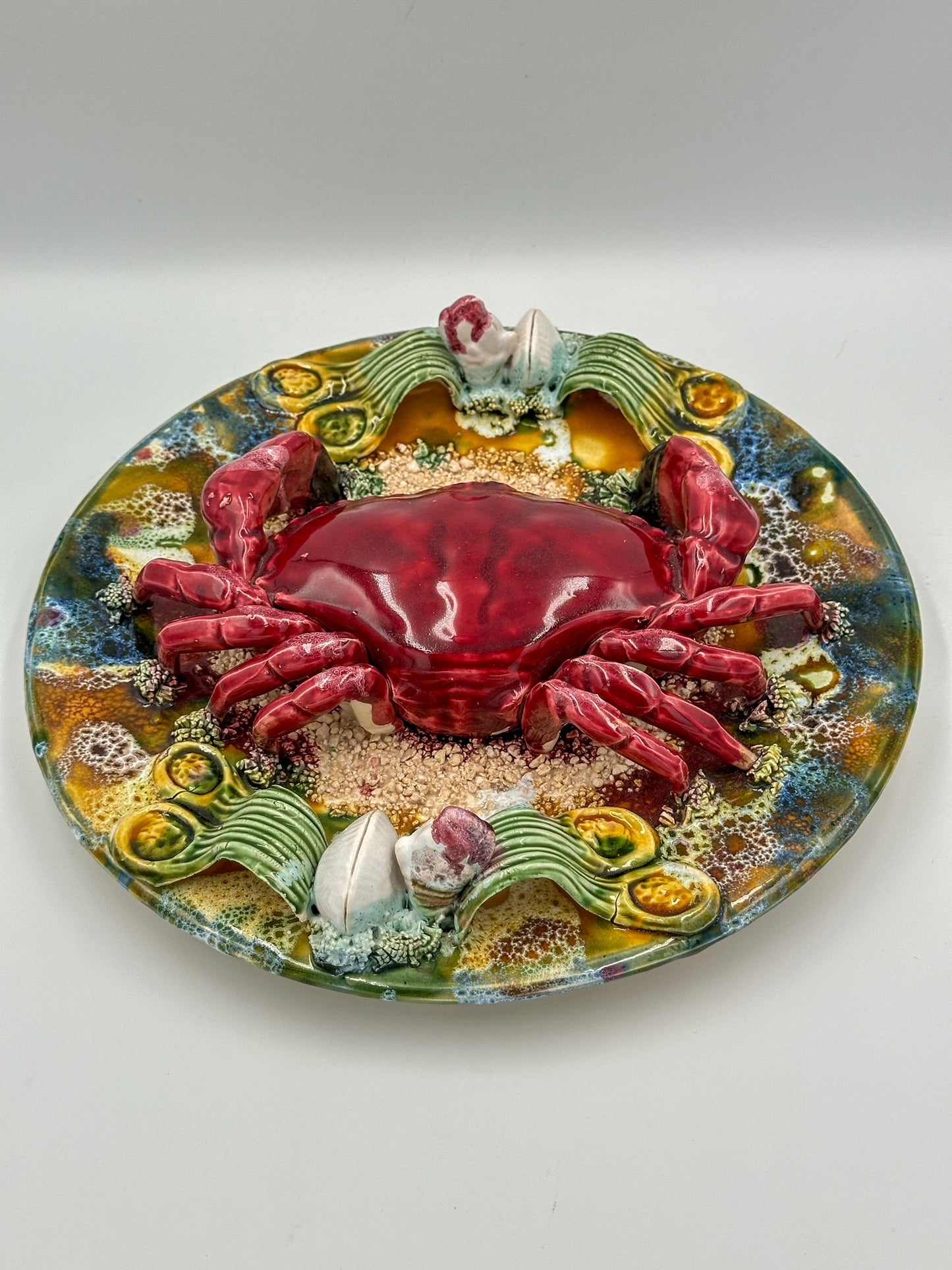 A decorative majolica display plate featuring a realistic depiction of a red crab with a detailed shell, surrounded by sand and shells, on a colorful background with floral and marine motifs.