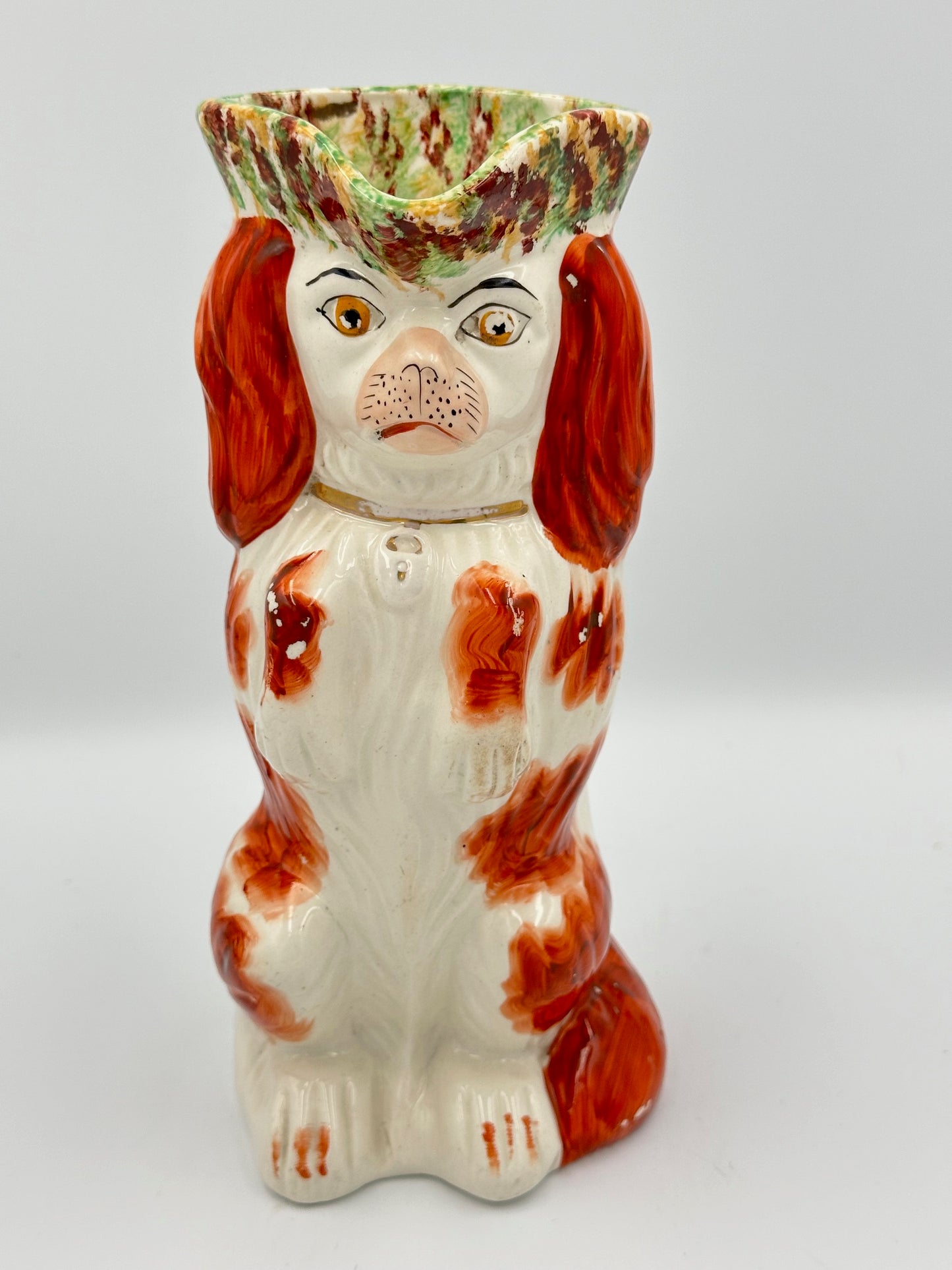 A ceramic pitcher shaped like a Staffordshire spaniel with a red and white coat, wearing a gold collar, and a green, yellow, and brown hat.