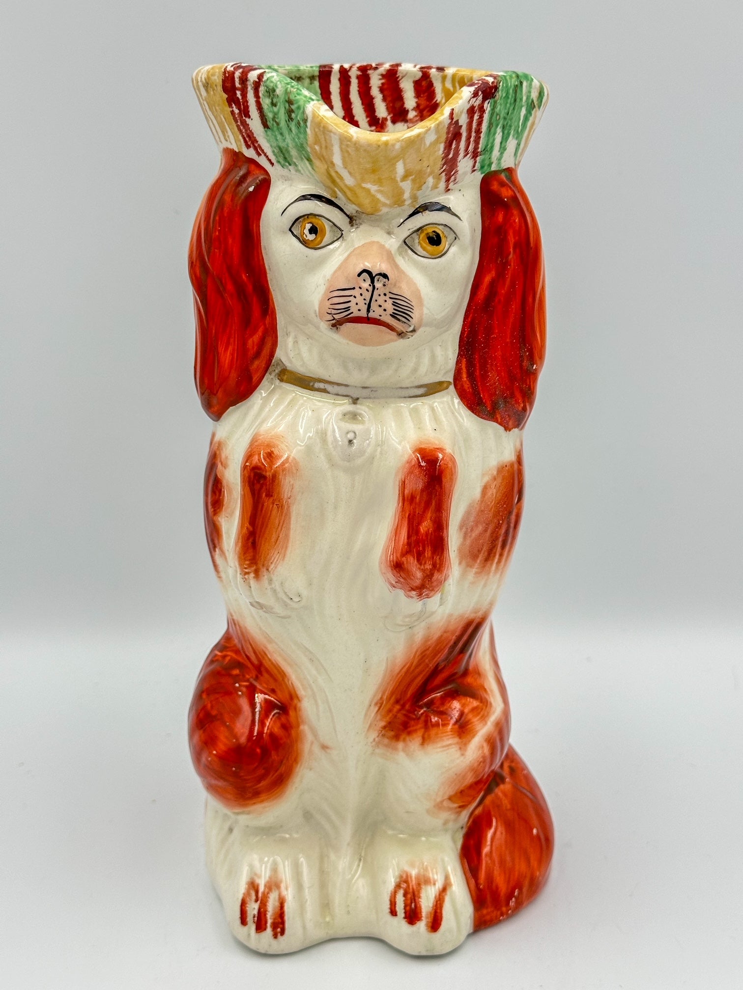 Antique 19th century Staffordshire ceramic pitcher in the shape of a King Charles Spaniel, featuring a begging pose with the dog's paws on its chest. The pitcher is primarily white with red and green accents.