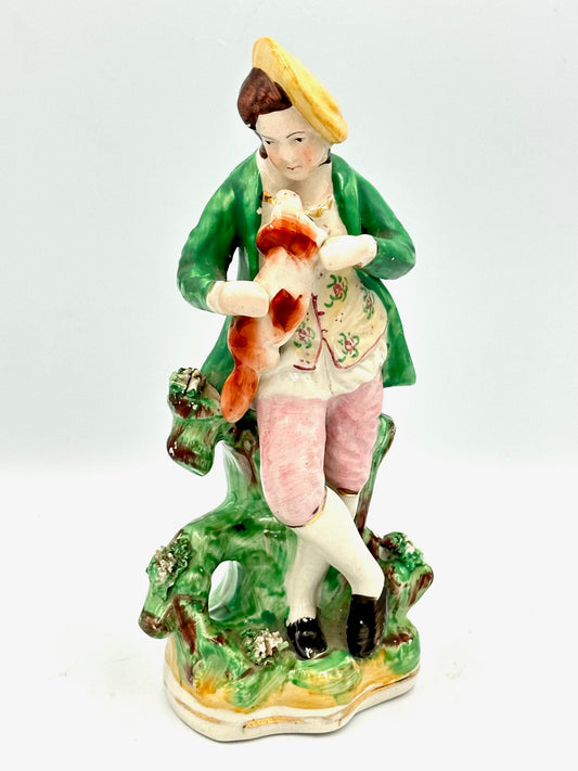 A flatback figurine depicting a well-dressed dandy holding a small dog in a garden setting. The dandy is wearing a green coat, white stockings, and black shoes, and the dog is red/russet in color.