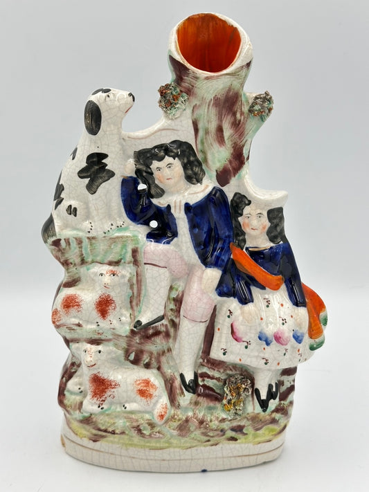 A ceramic spill vase flatback featuring a boy, girl, dog, and two lambs with a tree that has a hollow center for holding spills.