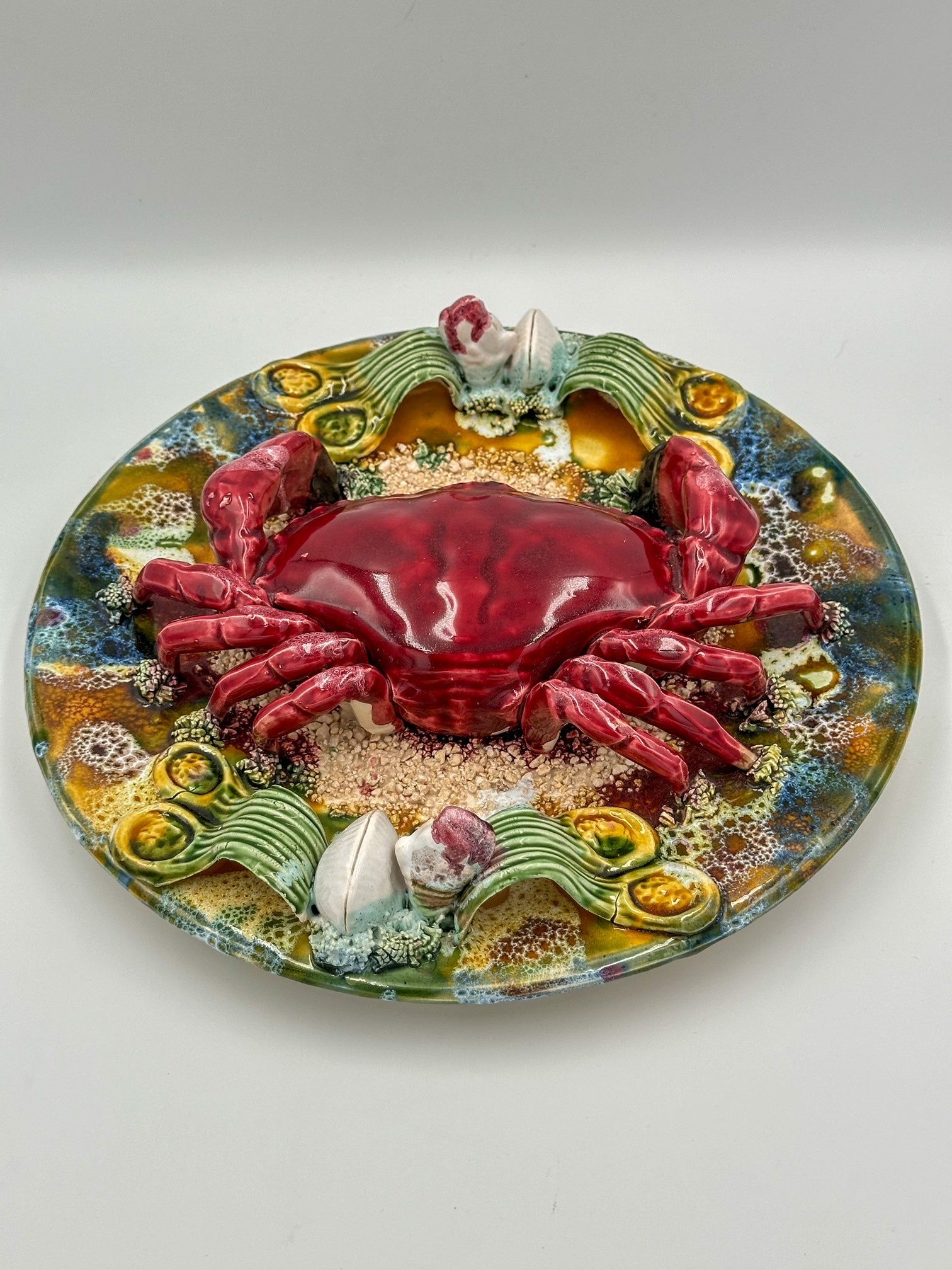 A decorative majolica display plate featuring a realistic depiction of a red crab with a detailed shell, surrounded by sand and shells, on a colorful background with floral and marine motifs.