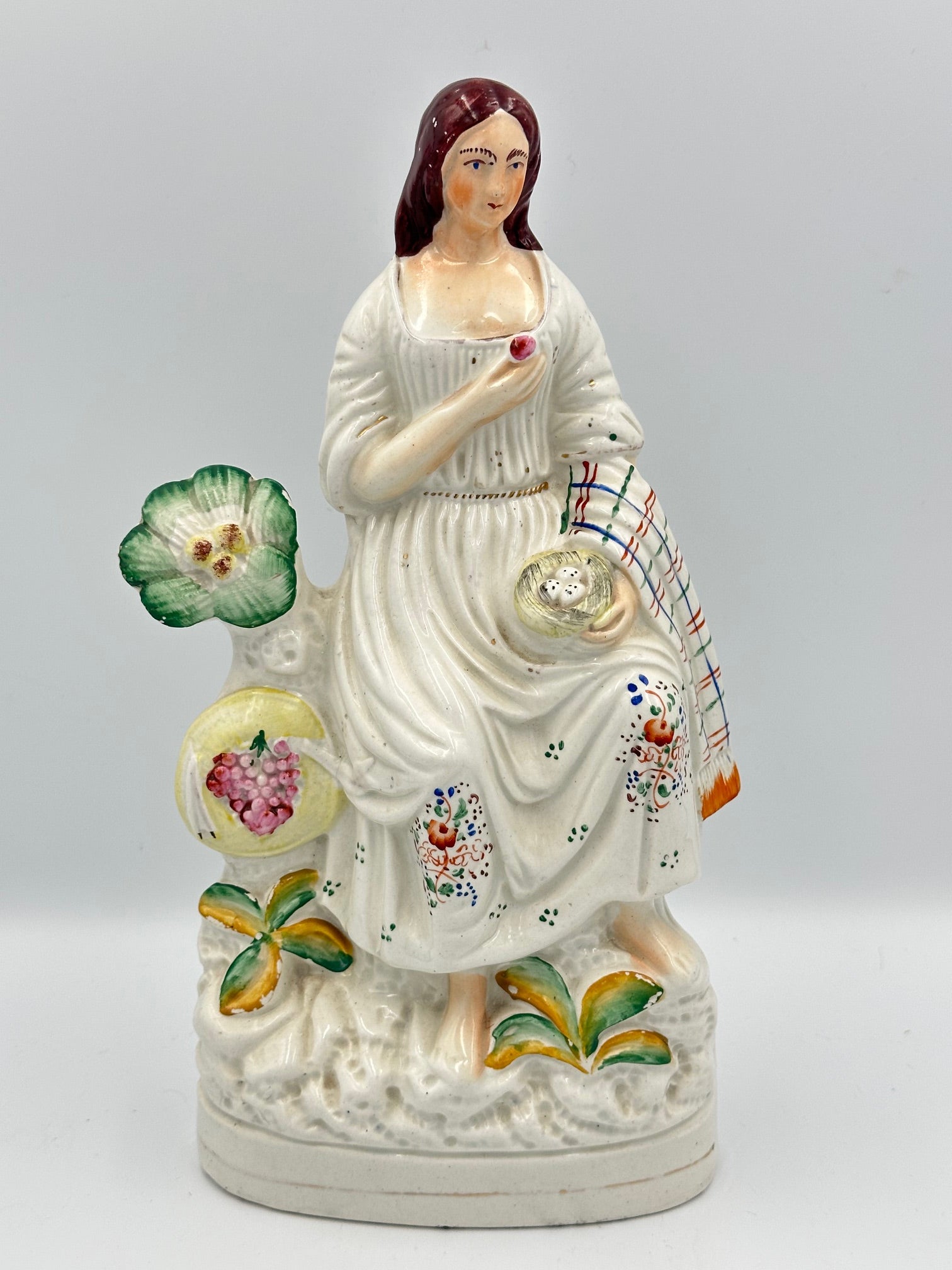 A white porcelain figurine of a woman pigeon fancier, holding a basket of eggs and a grape, with a colorful shawl draped over her left arm, set against a decorative backdrop of plants.