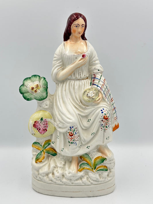 A white porcelain figurine of a woman pigeon fancier, holding a basket of eggs and a grape, with a colorful shawl draped over her left arm, set against a decorative backdrop of plants.