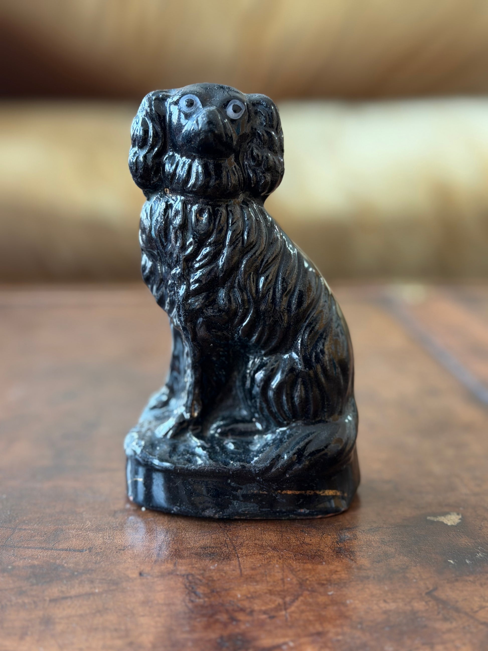 A small black dog figurine with detailed fur carving, sitting on a polished wooden surface with a softly blurred warm-toned background.