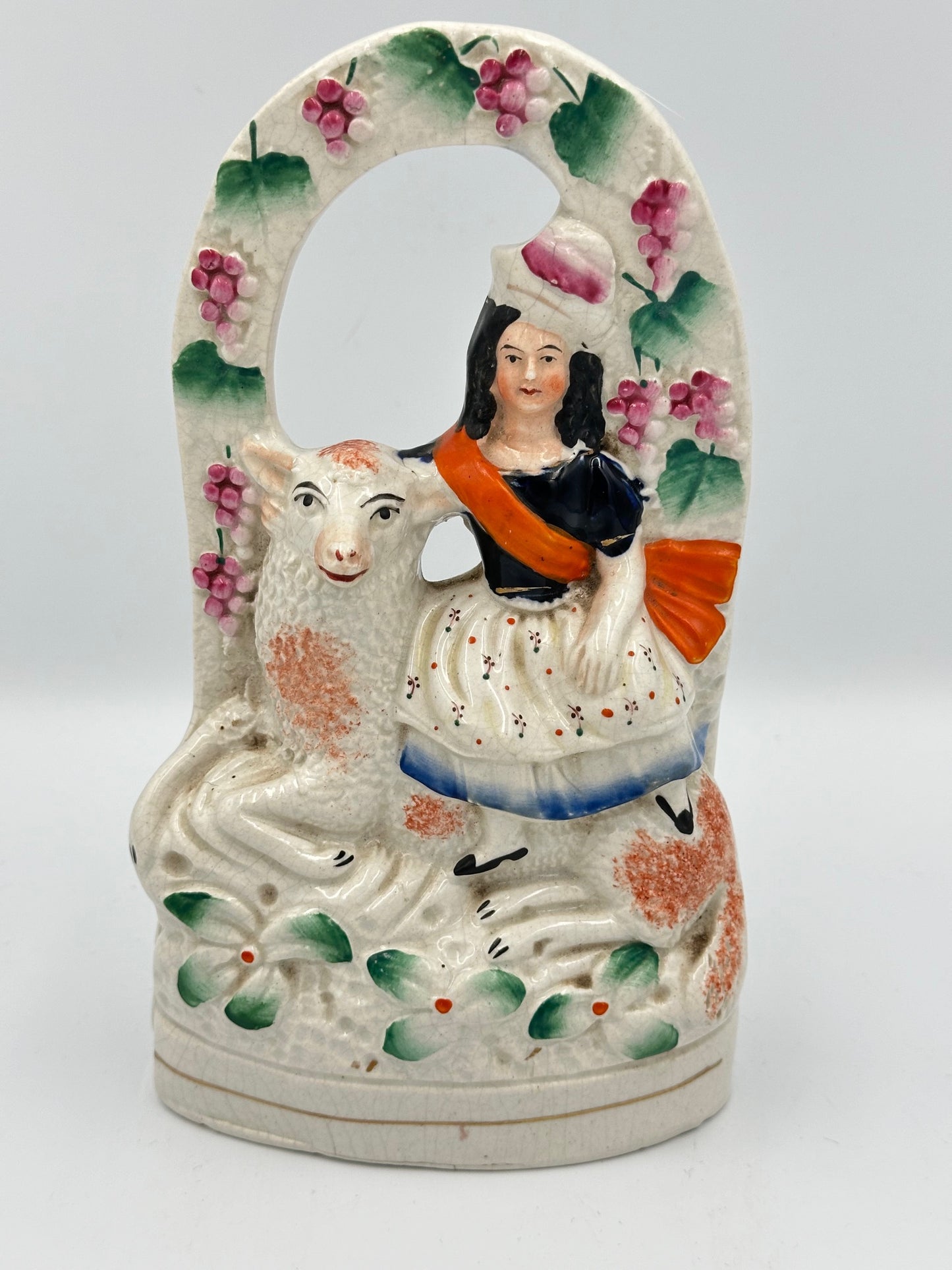 A ceramic flatback figure featuring a woman sitting on a sheep, with a grapevine and arbor in the background, adorned with floral patterns and a red sash.