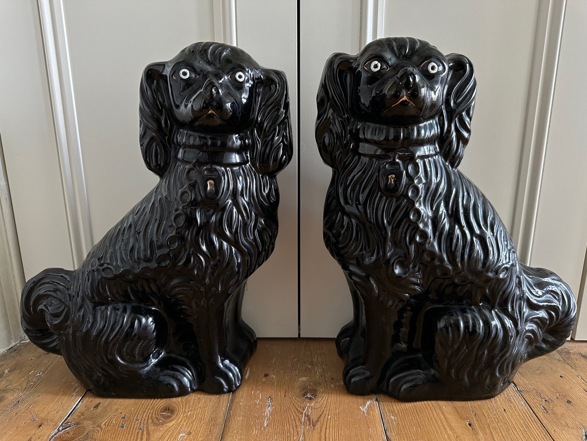 A pair of large black antique Staffordshire King Charles Spaniel dogs with white hand-painted eyes, featuring some gilding on collars, locks, and chains.