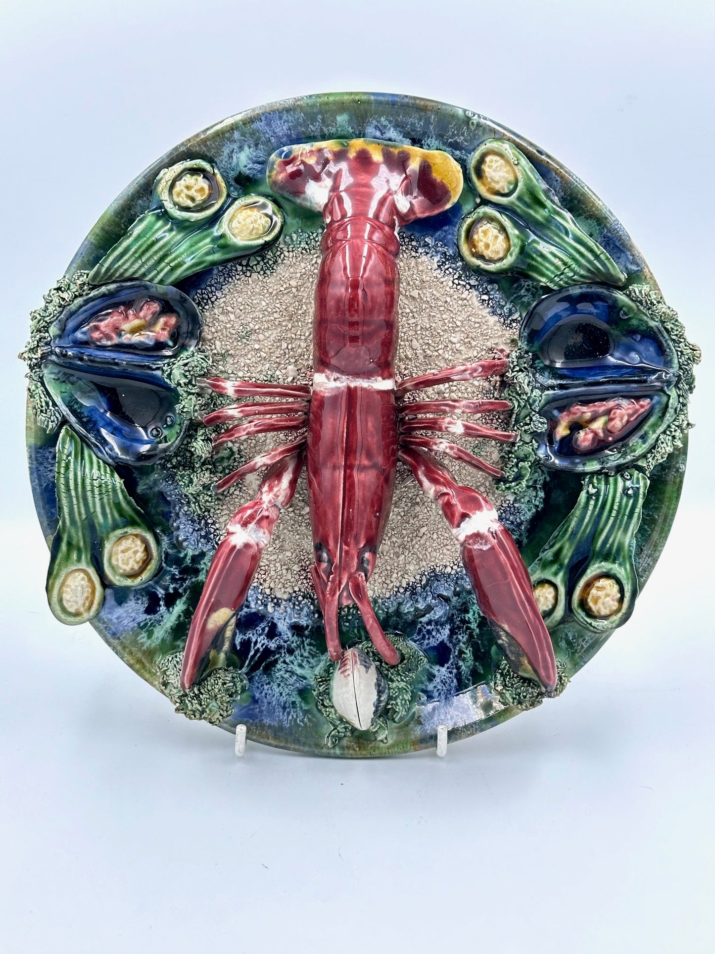 A decorative majolica plate featuring a central lobster with a detailed shell, surrounded by sand and flanked by two decorative elements, all on a white background. The plate has a vivid colorful border.