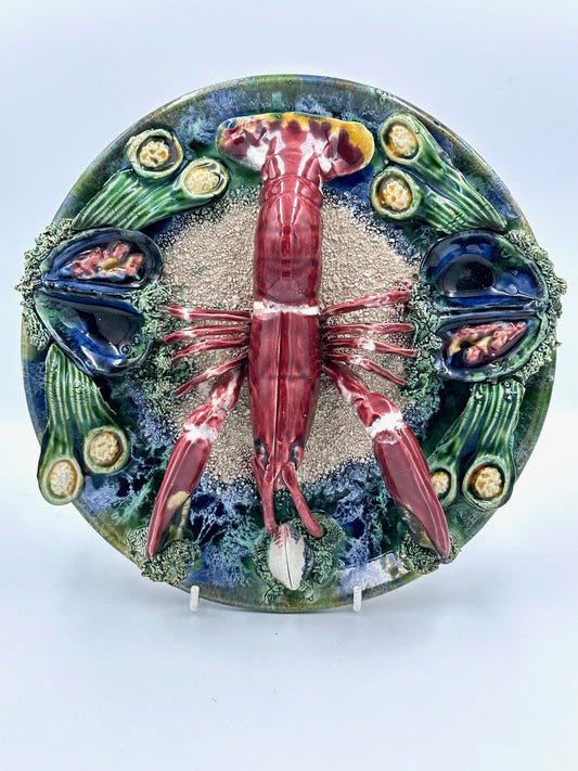 A decorative majolica plate featuring a central lobster with a detailed shell, surrounded by sand and flanked by two decorative elements, all on a white background. The plate has a vivid colorful border.