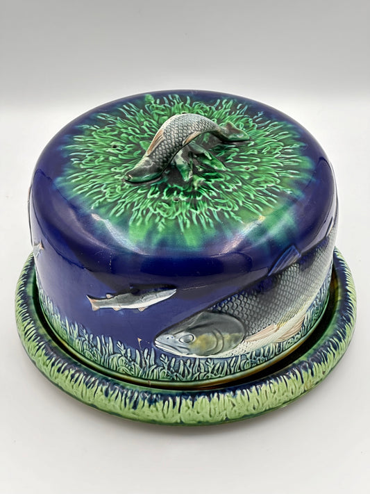 A vintage English Majolica cheese dome with a hand painted fish relief and green foilage on a cobalt blue background, featuring a raised fish handle on the top and a tortoise shell glazing technique on the bottom.