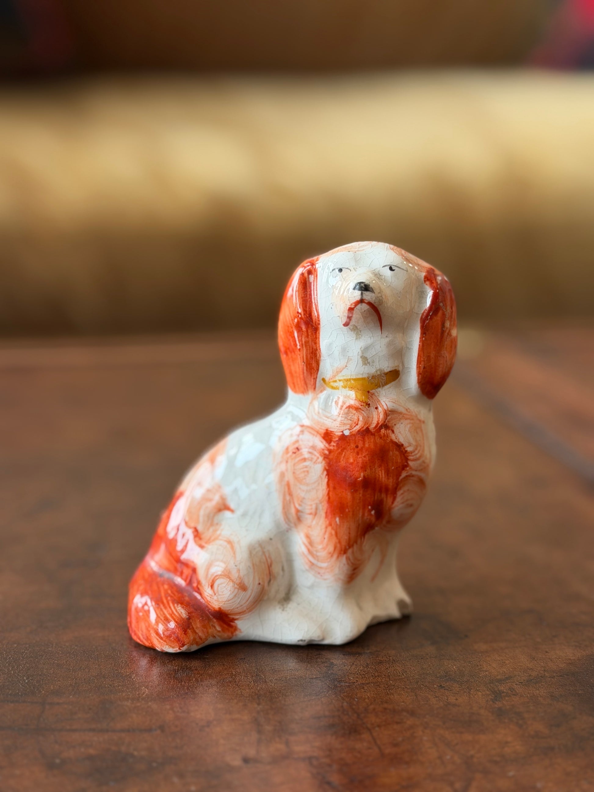 A small ceramic dog figurine with a white body and orange marbled patches, sitting on a wooden surface with a blurred indoor background.