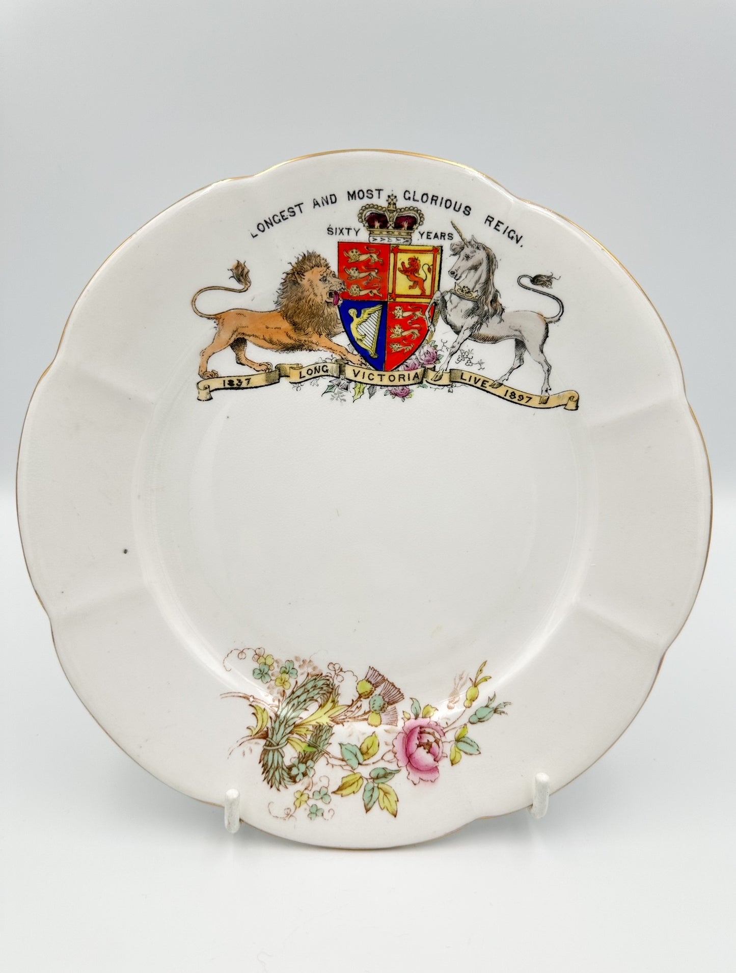 A white porcelain plate with a floral pattern and a commemorative design featuring a lion and unicorn, royal crest, and the words 'LONGSTOOD ON THE THRONE OF ENGLAND SIXTY YEARS'.