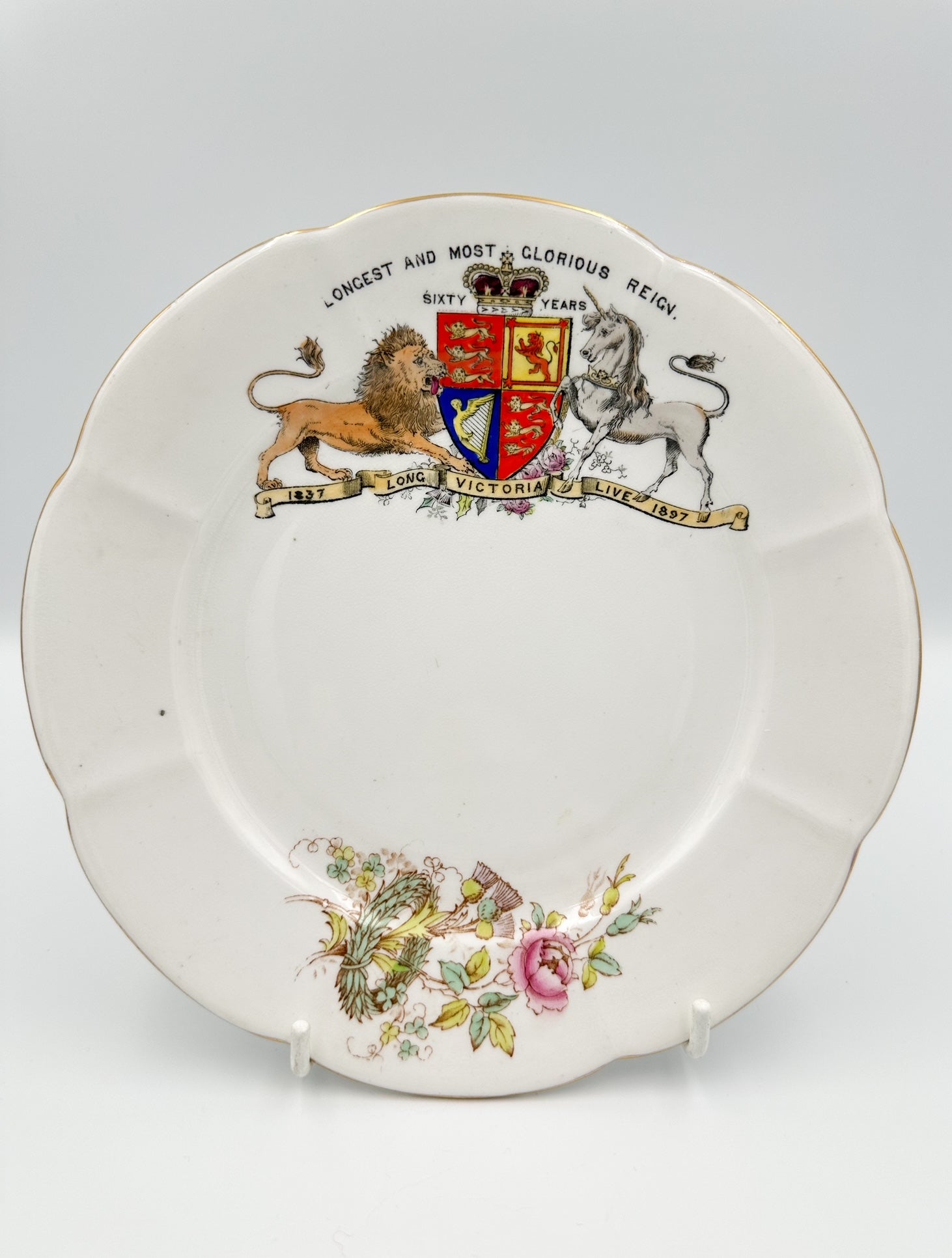 A white porcelain plate with a floral pattern and a commemorative design featuring a lion and unicorn, royal crest, and the words 'LONGSTOOD ON THE THRONE OF ENGLAND SIXTY YEARS'.