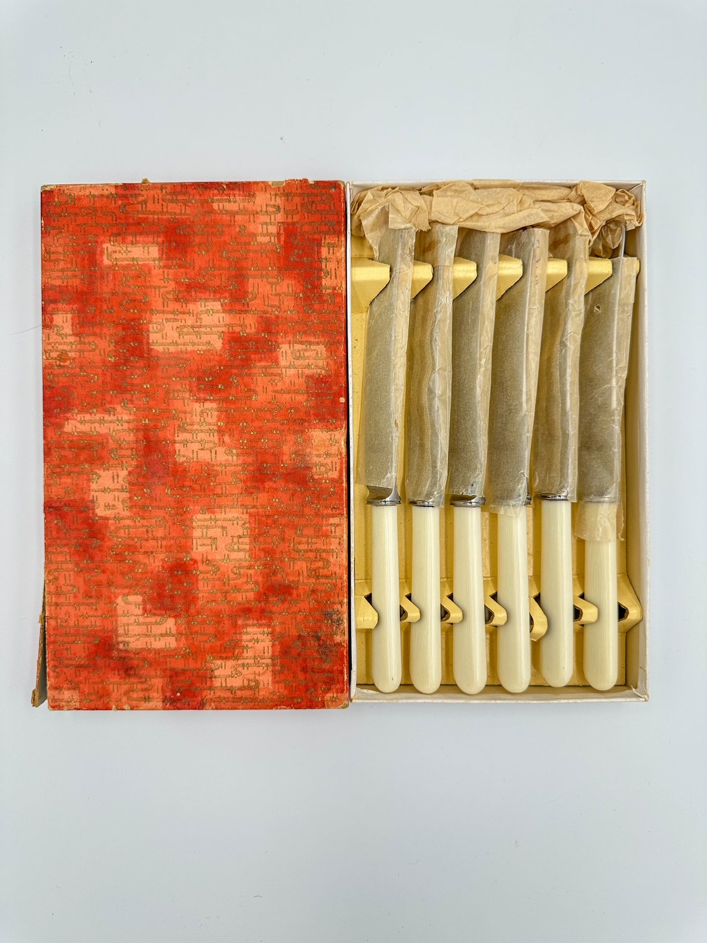 A set of six vintage stainless steel and Bakelite dinner knives, still wrapped in waxed paper, displayed in an open box with an orange and white patterned interior.