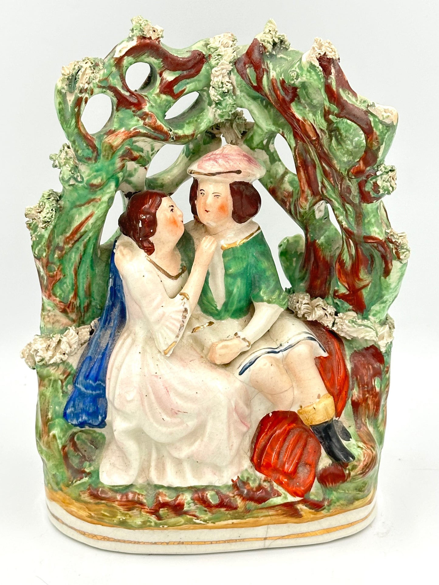 Staffordshire ceramic flatback figure depicting Romeo and Juliet, with green and blue cloaks, sitting on a bench under a tree.