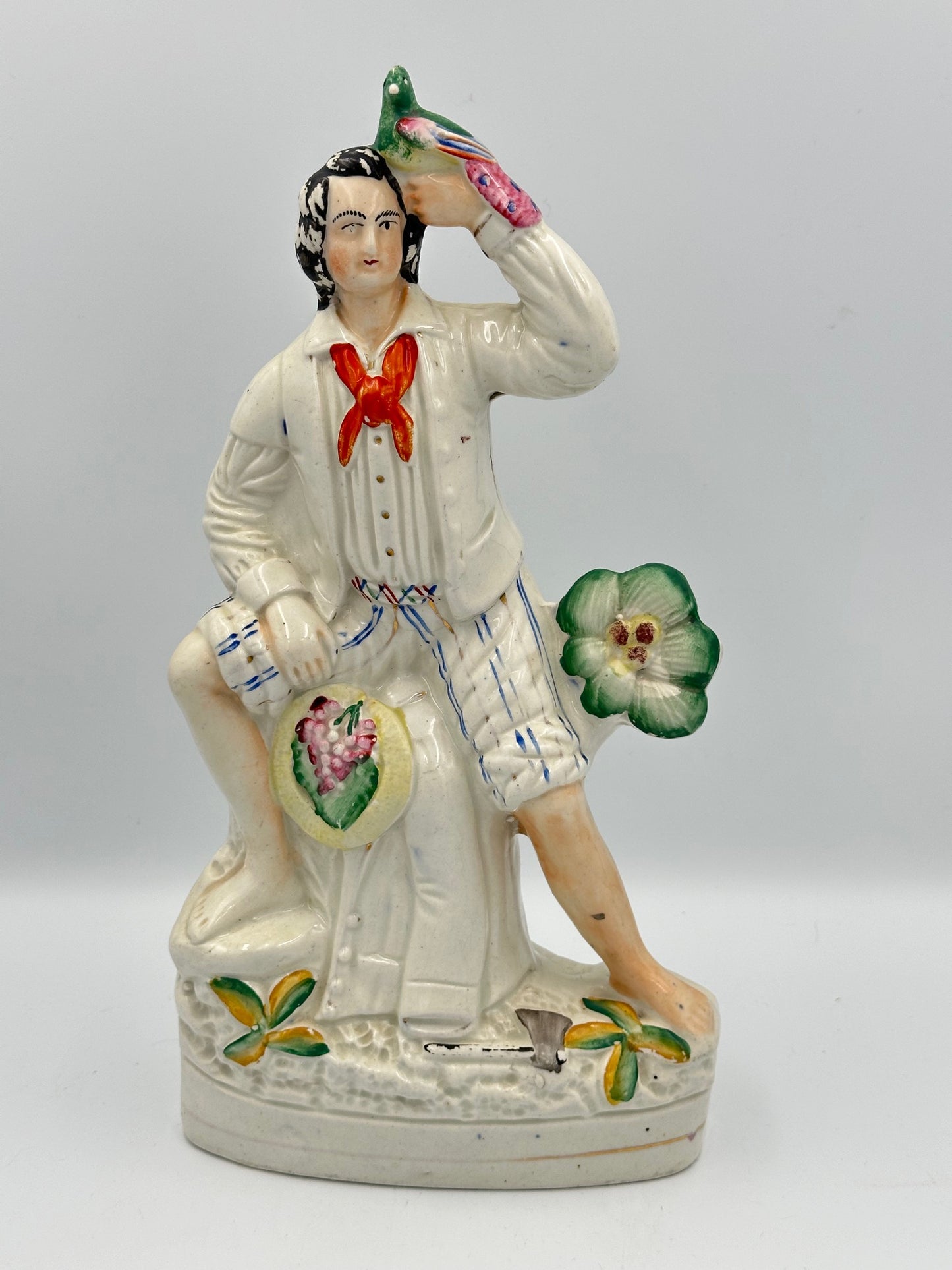 A ceramic flatback figurine of a 19th-century pigeon fancier, dressed in a white jacket with a red collar, blue striped trousers, and holding a pigeon on his head. Decorated with a bunch of grapes, plants, and an axe.