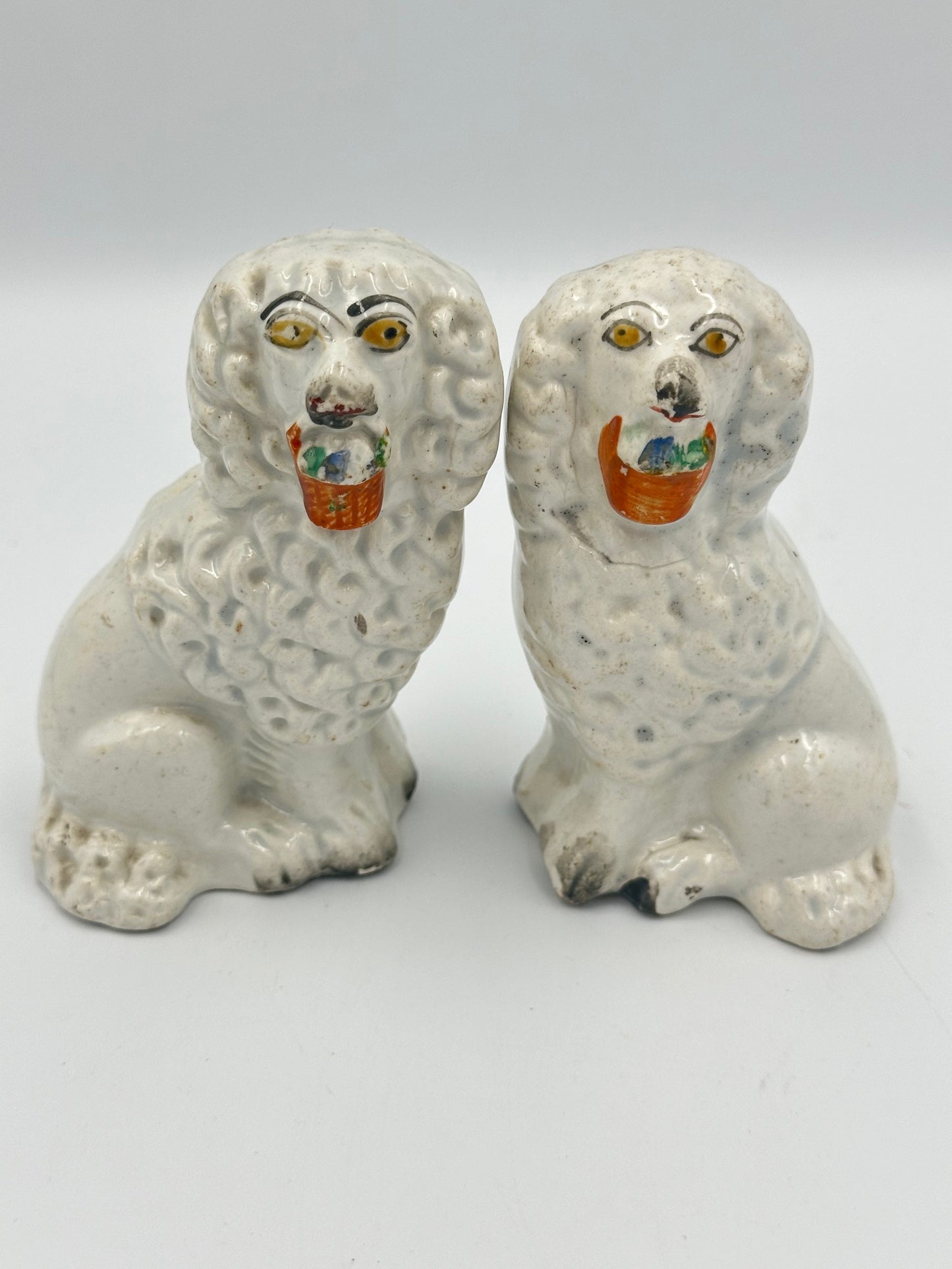 A pair of Staffordshire ceramic poodle figurines, each with a colorful basket in their mouth, seated facing forward.