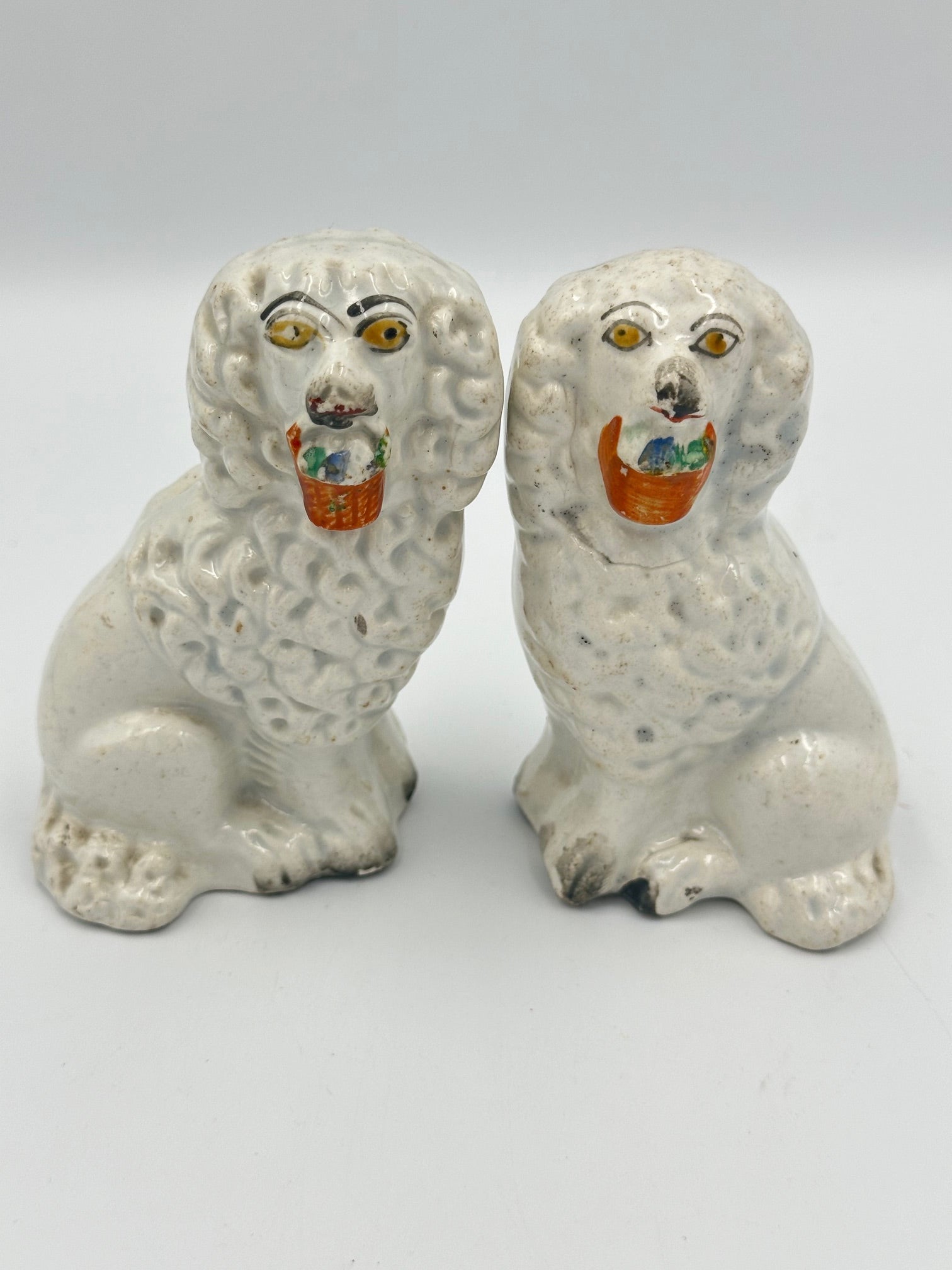 A pair of Staffordshire ceramic poodle figurines, each with a colorful basket in their mouth, seated facing forward.