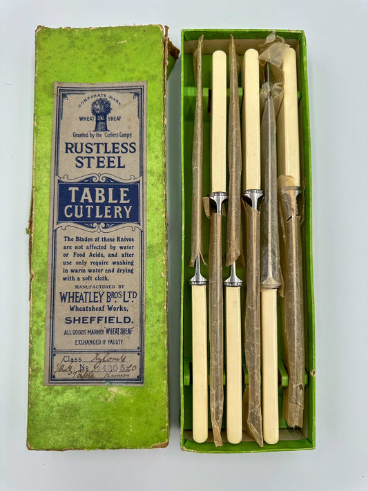 A set of six vintage stainless steel and Bakelite dinner knives still wrapped in waxed paper, displayed in an open box with a label reading 'RUSTLESS STEEL TABLE CUTLERY'.