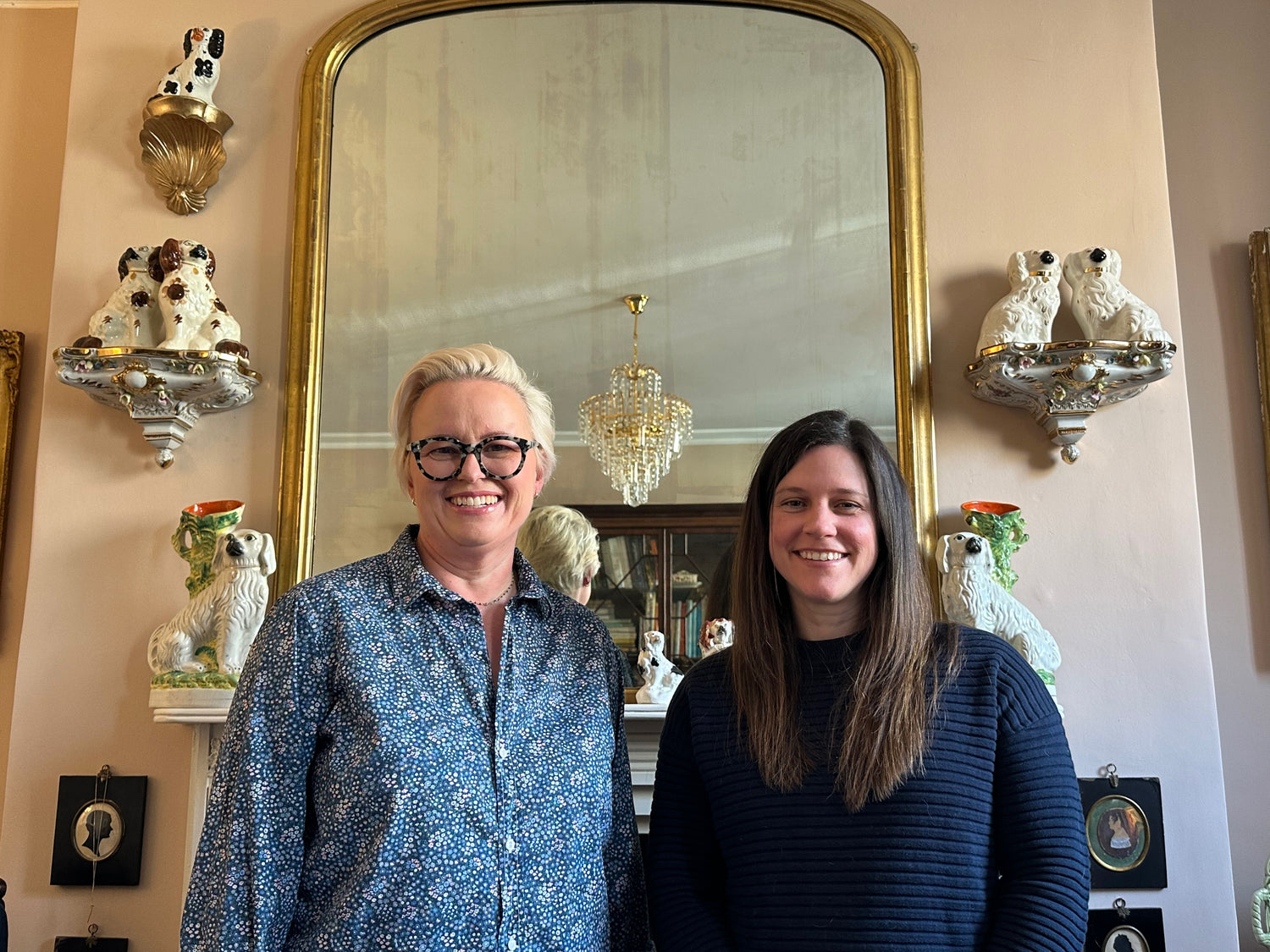 Two women standing in a room with decorative wall mirrors and animal figurines.